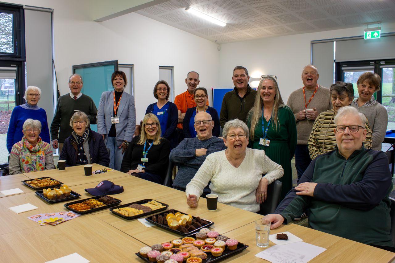 A group of around 15 people sitting and standing around a table. Some of them are wearing scrubs, they are smiling and laughing together. On the table in front of them is a selection of cakes and drinks.