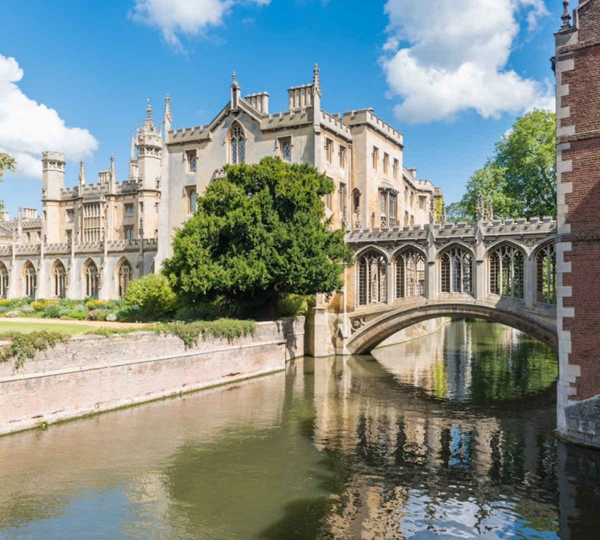  A historic stone bridge with gothic-style windows, known as the Bridge of Sighs, spans a calm river between two ornate university buildings in Cambridge, England. Lush greenery and a clear blue sky complete the picturesque scene.