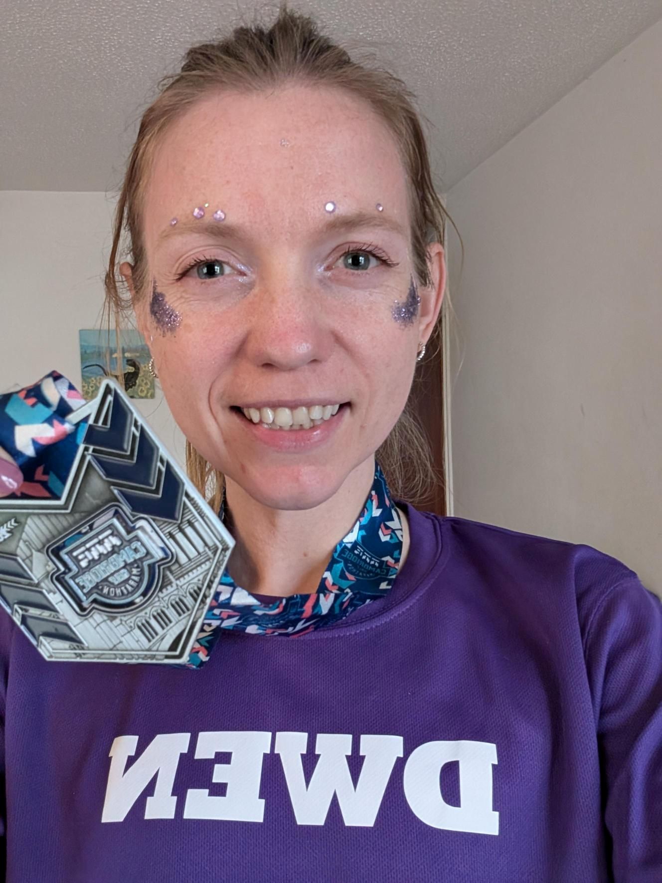 Image shows Ceiridwen Walker after the Cambridge Half Marathon. She is wearing a shirt in the signature my wish charity purple, and has purple glitter and gems on her face. She is holding up a medal to her face, and she is smiling at the camera.