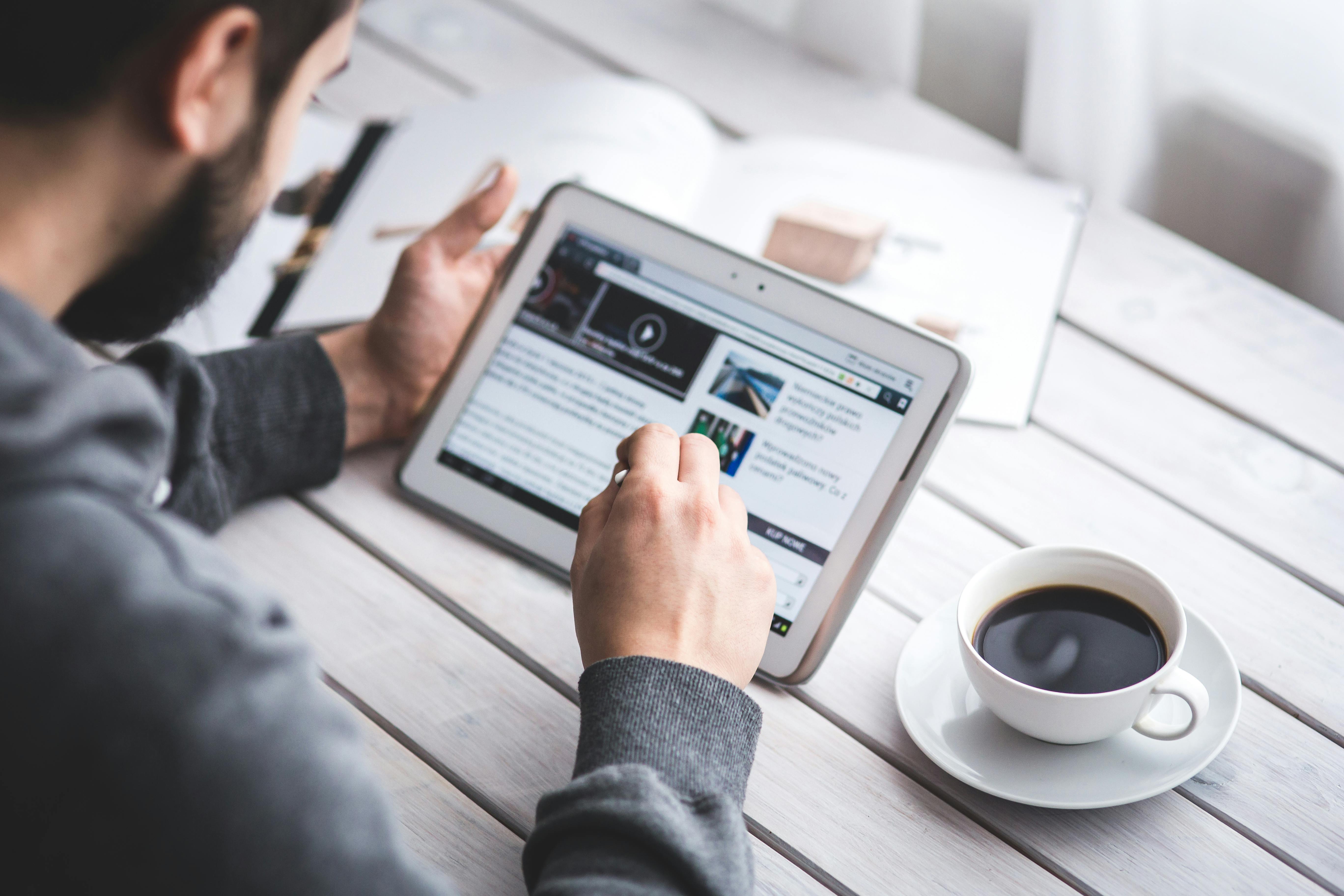 Man using a tablet with a stylus while reading online content, sitting at a wooden table with a cup of black coffee and an open notebook in the background.