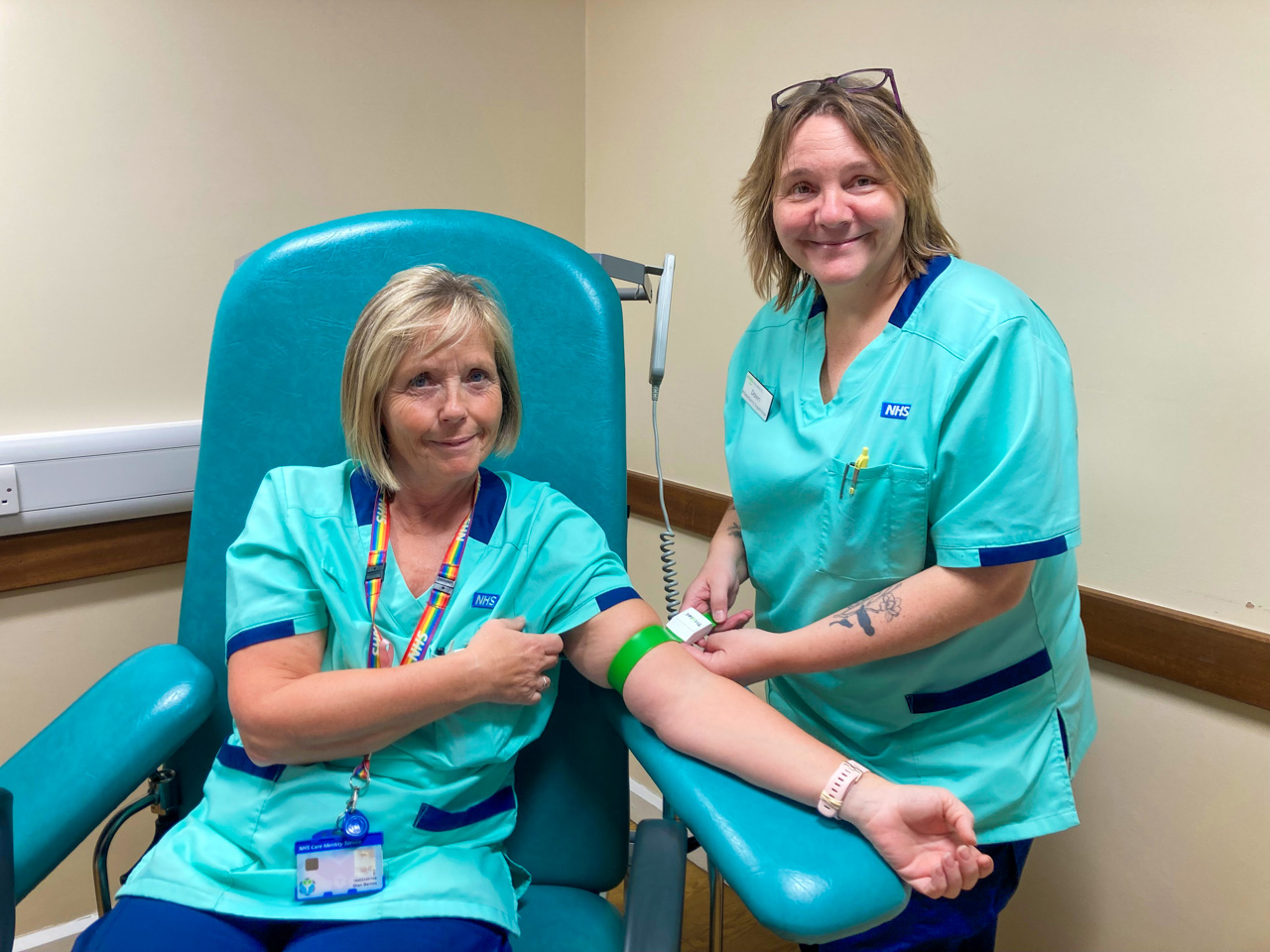 One woman, standing, attaching a tourniquet to the arm of a woman sat down in a chair