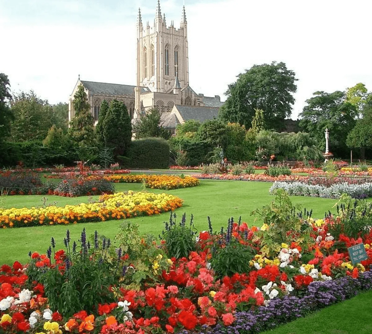 A vibrant formal garden filled with colorful flowerbeds, featuring red, yellow, purple, and white blooms, set against manicured lawns. In the background stands the historic St Edmundsbury Cathedral in Bury St Edmunds, surrounded by trees and greenery.